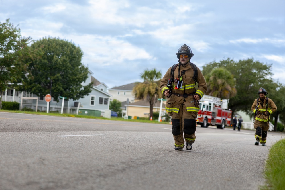 Two firefighters in full turnout gear are walking along a residential street toward the camera. A red fire truck is visible in the background, suggesting an active emergency response or post-incident scene. The setting appears to be a suburban neighborhood with palm trees and residential homes.