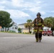 Two firefighters in full turnout gear are walking along a residential street toward the camera. A red fire truck is visible in the background, suggesting an active emergency response or post-incident scene. The setting appears to be a suburban neighborhood with palm trees and residential homes.