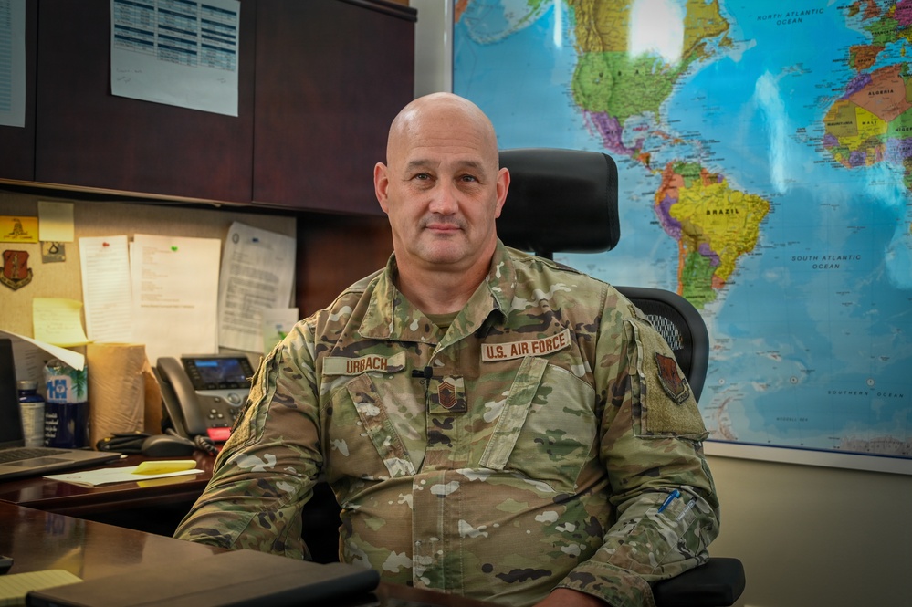 A U.S. Air Force senior enlisted member named Urbach is seated at a desk in what appears to be a military office setting. He is wearing Operational Camouflage Pattern (OCP) uniform with a Chief Master Sergeant rank insignia visible on his chest. A large world map is displayed prominently on the wall behind him.
