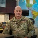 A U.S. Air Force senior enlisted member named Urbach is seated at a desk in what appears to be a military office setting. He is wearing Operational Camouflage Pattern (OCP) uniform with a Chief Master Sergeant rank insignia visible on his chest. A large world map is displayed prominently on the wall behind him.