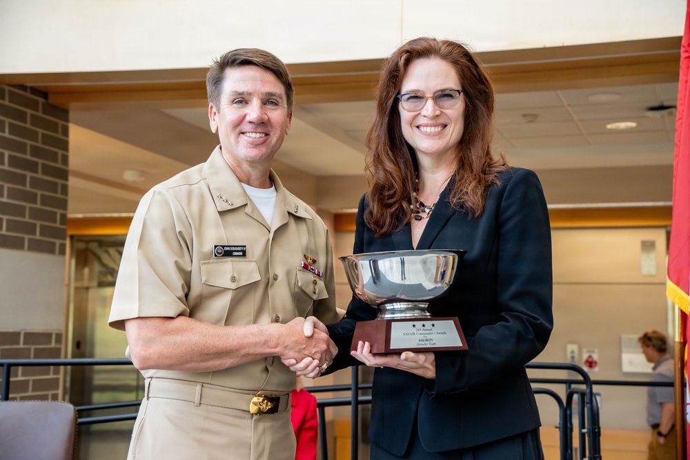 A U.S. Navy officer in tan service uniform is presenting a silver trophy bowl to a woman in a black blazer during what appears to be a formal awards ceremony. The trophy is mounted on a wooden base with a plaque that references 'NAVAIR Commander's Awards.' Both individuals are smiling and shaking hands while jointly holding the award.