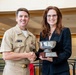 A U.S. Navy officer in tan service uniform is presenting a silver trophy bowl to a woman in a black blazer during what appears to be a formal awards ceremony. The trophy is mounted on a wooden base with a plaque that references 'NAVAIR Commander's Awards.' Both individuals are smiling and shaking hands while jointly holding the award.
