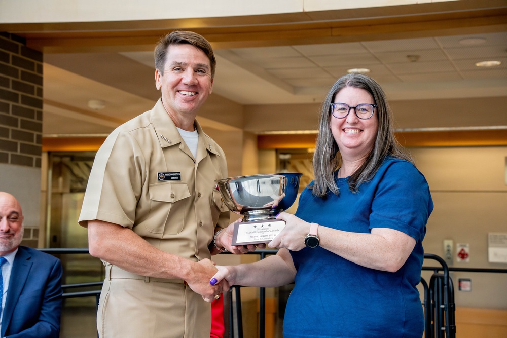 A U.S. Navy officer in tan service uniform is presenting a silver bowl trophy to a civilian woman wearing a blue top and glasses. Both individuals are smiling and shaking hands while jointly holding the award. The setting appears to be an indoor facility, likely during a formal awards ceremony.