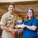 A U.S. Navy officer in tan service uniform is presenting a silver bowl trophy to a civilian woman wearing a blue top and glasses. Both individuals are smiling and shaking hands while jointly holding the award. The setting appears to be an indoor facility, likely during a formal awards ceremony.