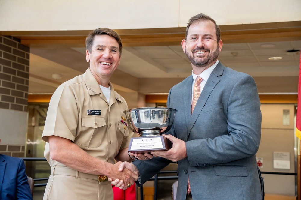 A U.S. Navy officer in tan service uniform and a civilian in a gray suit are jointly holding a silver trophy bowl mounted on a wooden base with a plaque. The two men are shaking hands and smiling, suggesting a formal award or recognition ceremony. The setting appears to be an indoor institutional facility.