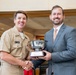 A U.S. Navy officer in tan service uniform and a civilian in a gray suit are jointly holding a silver trophy bowl mounted on a wooden base with a plaque. The two men are shaking hands and smiling, suggesting a formal award or recognition ceremony. The setting appears to be an indoor institutional facility.