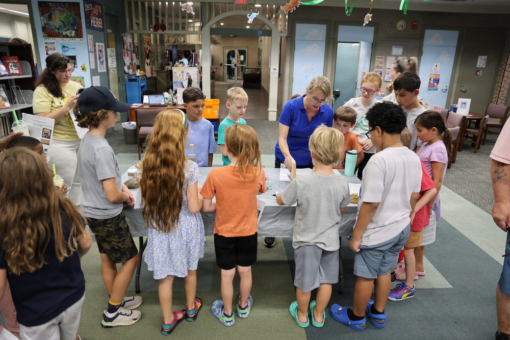 A group of children gathered around a table in what appears to be a public library, participating in a hands-on educational activity led by an adult in a blue polo shirt. The setting is decorated with hanging paper animals and colorful streamers, suggesting a themed summer reading program or special event. Several adults, including parents and staff, are present supervising the activity.