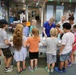 A group of children gathered around a table in what appears to be a public library, participating in a hands-on educational activity led by an adult in a blue polo shirt. The setting is decorated with hanging paper animals and colorful streamers, suggesting a themed summer reading program or special event. Several adults, including parents and staff, are present supervising the activity.