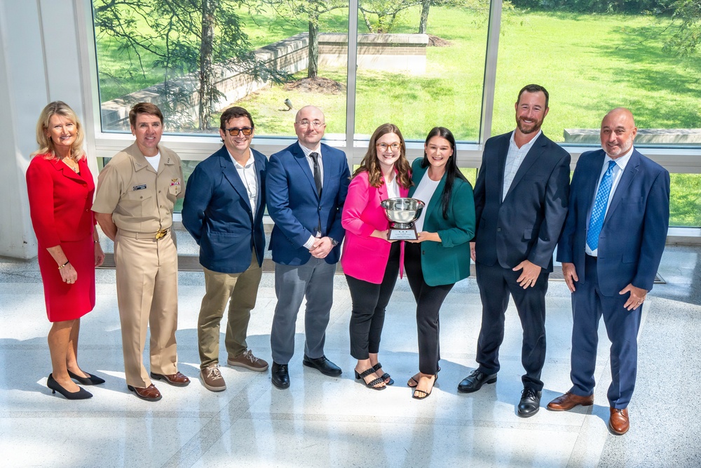 A group of eight individuals pose for a formal group photograph in what appears to be a modern building with large windows overlooking a green outdoor area. Two women in the center are jointly holding a trophy or award bowl, suggesting a recognition or award ceremony. The group includes one person in a military uniform and several others in professional business attire.