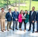 A group of eight individuals pose for a formal group photograph in what appears to be a modern building with large windows overlooking a green outdoor area. Two women in the center are jointly holding a trophy or award bowl, suggesting a recognition or award ceremony. The group includes one person in a military uniform and several others in professional business attire.