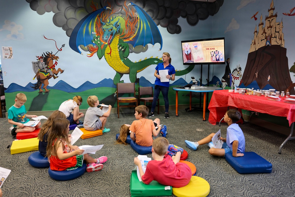 An adult instructor in blue scrubs leads a children's educational program in what appears to be a library or community center reading room. Approximately 10 children are seated on colorful floor cushions facing a TV screen displaying 'THE END' from a storybook presentation. The room features a vibrant fantasy-themed mural depicting a dragon, knight on horseback, and castle.
