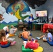 An adult instructor in blue scrubs leads a children's educational program in what appears to be a library or community center reading room. Approximately 10 children are seated on colorful floor cushions facing a TV screen displaying 'THE END' from a storybook presentation. The room features a vibrant fantasy-themed mural depicting a dragon, knight on horseback, and castle.