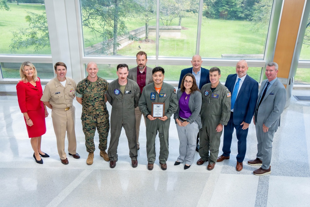 A group of approximately ten individuals pose for a formal group photograph in what appears to be a modern building lobby with large windows overlooking a green landscape. The group includes military personnel in various uniforms (Navy, Marine Corps, and flight suits) alongside civilians in business attire. A central figure in a flight suit is holding a plaque or award, suggesting this is an award or recognition ceremony.