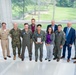 A group of approximately ten individuals pose for a formal group photograph in what appears to be a modern building lobby with large windows overlooking a green landscape. The group includes military personnel in various uniforms (Navy, Marine Corps, and flight suits) alongside civilians in business attire. A central figure in a flight suit is holding a plaque or award, suggesting this is an award or recognition ceremony.