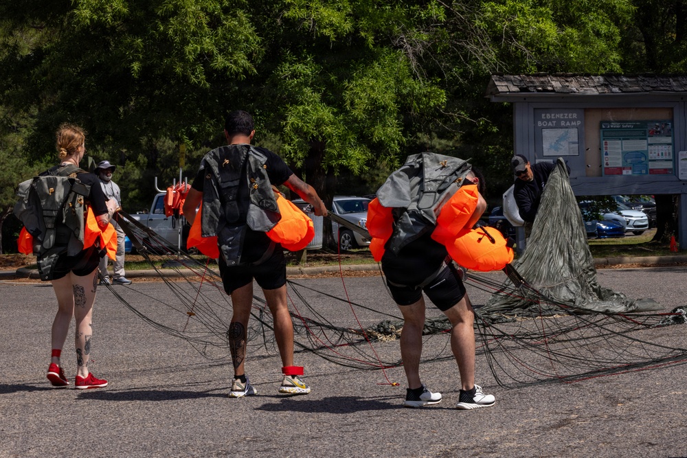 Three individuals wearing black athletic clothing and parachute rigs with bright orange deployment bags are handling parachute equipment in a parking lot near the 'Ebenezer Boat Ramp.' The parachute canopy and lines are spread across the asphalt, suggesting the group is packing or inspecting gear after a jump. A fourth person in the background appears to be assisting with the parachute canopy.