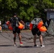 Three individuals wearing black athletic clothing and parachute rigs with bright orange deployment bags are handling parachute equipment in a parking lot near the 'Ebenezer Boat Ramp.' The parachute canopy and lines are spread across the asphalt, suggesting the group is packing or inspecting gear after a jump. A fourth person in the background appears to be assisting with the parachute canopy.