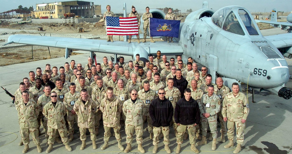 A large group of military personnel pose for a unit photo in front of an A-10 Thunderbolt II (Warthog) attack aircraft bearing the tail number 659. Four individuals standing on the aircraft's wing hold an American flag and what appears to be a Pennsylvania state flag. The setting appears to be a forward operating base in a desert environment, likely during Operation Iraqi Freedom or Operation Enduring Freedom.