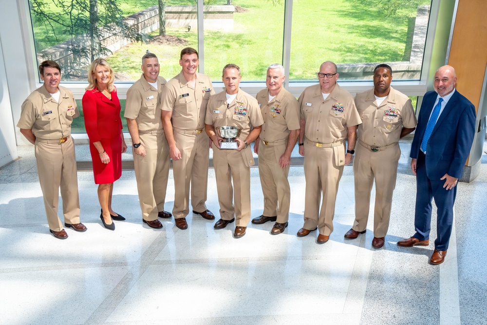 A group of nine individuals pose for a formal photograph in what appears to be a modern building lobby or atrium. Seven of the individuals are wearing U.S. Navy tan/khaki service uniforms, one woman is dressed in a red business suit, and one man is wearing a civilian navy blue suit. The central figure is holding a silver trophy or award bowl, suggesting this is an award or recognition ceremony.