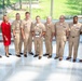 A group of nine individuals pose for a formal photograph in what appears to be a modern building lobby or atrium. Seven of the individuals are wearing U.S. Navy tan/khaki service uniforms, one woman is dressed in a red business suit, and one man is wearing a civilian navy blue suit. The central figure is holding a silver trophy or award bowl, suggesting this is an award or recognition ceremony.