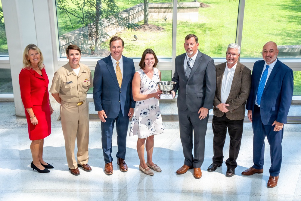 A group of seven adults pose for a formal photograph in what appears to be a modern building with large windows overlooking a green outdoor area. One individual in the center is holding a silver bowl trophy or award, suggesting this is an award presentation ceremony. The group includes one person in a U.S. military uniform (tan/khaki service dress) and several others in business attire.