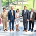 A group of seven adults pose for a formal photograph in what appears to be a modern building with large windows overlooking a green outdoor area. One individual in the center is holding a silver bowl trophy or award, suggesting this is an award presentation ceremony. The group includes one person in a U.S. military uniform (tan/khaki service dress) and several others in business attire.
