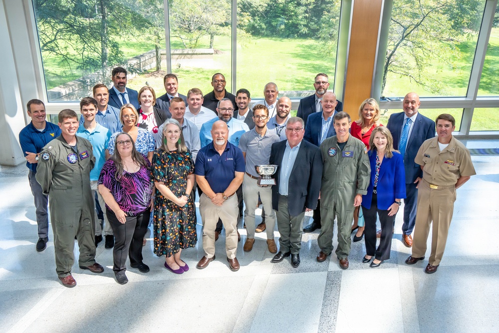 A large group of approximately 25-30 individuals poses for a formal group photograph inside a modern building with floor-to-ceiling windows overlooking a green landscape. The group includes individuals in military uniforms (flight suits and Navy dress uniform), business attire, and business casual clothing. One person in the front row is holding a silver trophy or bowl, suggesting an award ceremony or recognition event.