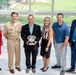 A group of six adults pose for a formal photograph in what appears to be a modern building lobby or atrium. The central figure holds a silver trophy bowl, suggesting an award ceremony or recognition event. One individual is wearing a U.S. Navy uniform, indicating a military-affiliated occasion.