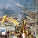 A U.S. military soldier in woodland camouflage BDUs stands guard on a debris-covered street in the foreground, while rescue and recovery workers operate heavy equipment amid massive structural rubble in the background. A yellow excavator labeled 'SUMMIT' is actively working through the debris pile, with smoke visible rising from the wreckage. Multiple personnel in hard hats and high-visibility vests are present, along with law enforcement vehicles.