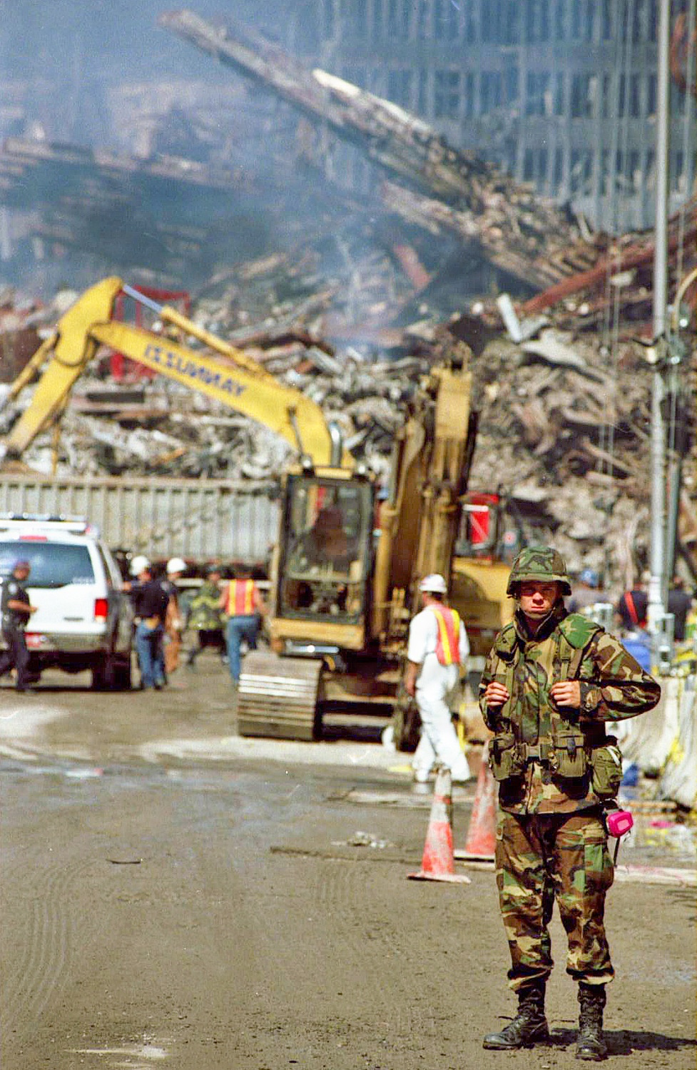 A U.S. military soldier in woodland camouflage BDUs stands guard on a debris-covered street in the foreground, while rescue and recovery workers operate heavy equipment amid massive structural rubble in the background. A yellow excavator labeled 'SUMMIT' is actively working through the debris pile, with smoke visible rising from the wreckage. Multiple personnel in hard hats and high-visibility vests are present, along with law enforcement vehicles.