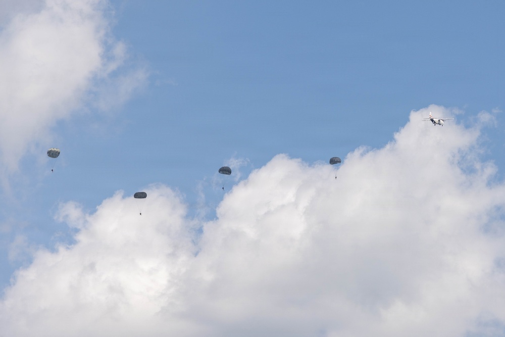 The image shows a military or airborne operation with multiple parachutists descending through a partly cloudy blue sky. An aircraft, likely the delivery platform, is visible in the upper right portion of the frame. The parachutes appear to be round military-style canopies, consistent with a mass tactical jump or airborne exercise.