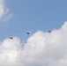 The image shows a military or airborne operation with multiple parachutists descending through a partly cloudy blue sky. An aircraft, likely the delivery platform, is visible in the upper right portion of the frame. The parachutes appear to be round military-style canopies, consistent with a mass tactical jump or airborne exercise.