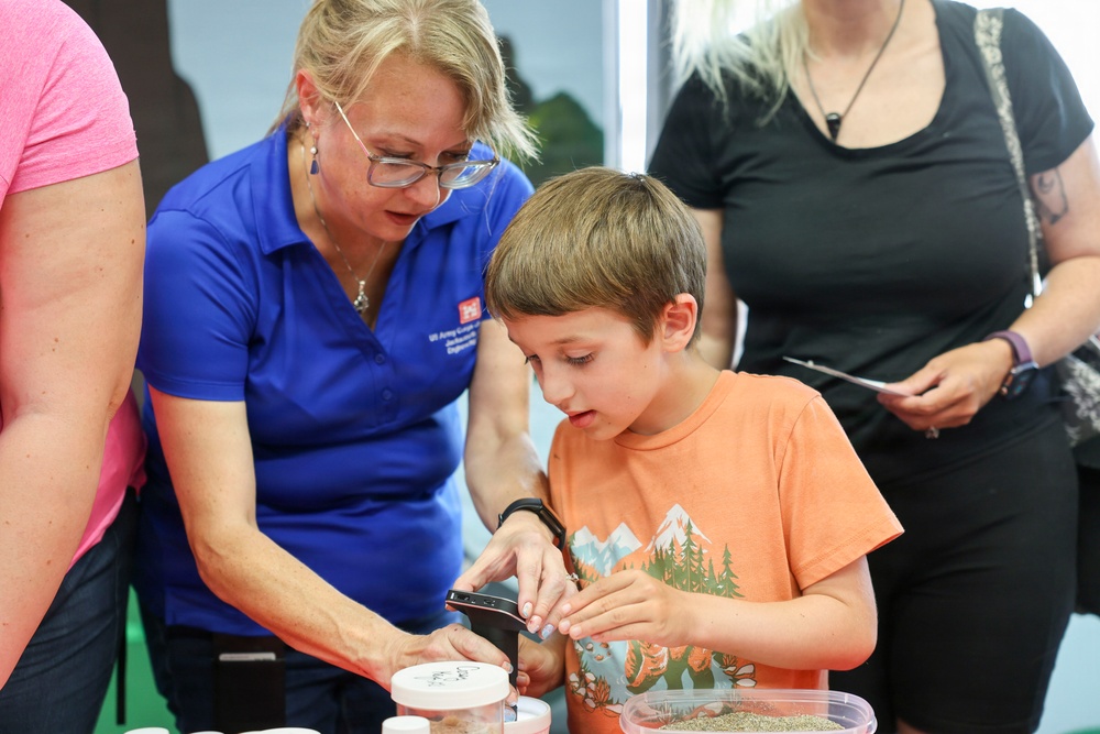 A woman in a blue polo shirt with a name badge is guiding a young boy through a hands-on activity at what appears to be an educational or science outreach event. The boy in an orange t-shirt is engaged and focused, examining something in a container using a handheld magnifier. Several adults are visible in the background observing the activity.