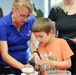 A woman in a blue polo shirt with a name badge is guiding a young boy through a hands-on activity at what appears to be an educational or science outreach event. The boy in an orange t-shirt is engaged and focused, examining something in a container using a handheld magnifier. Several adults are visible in the background observing the activity.