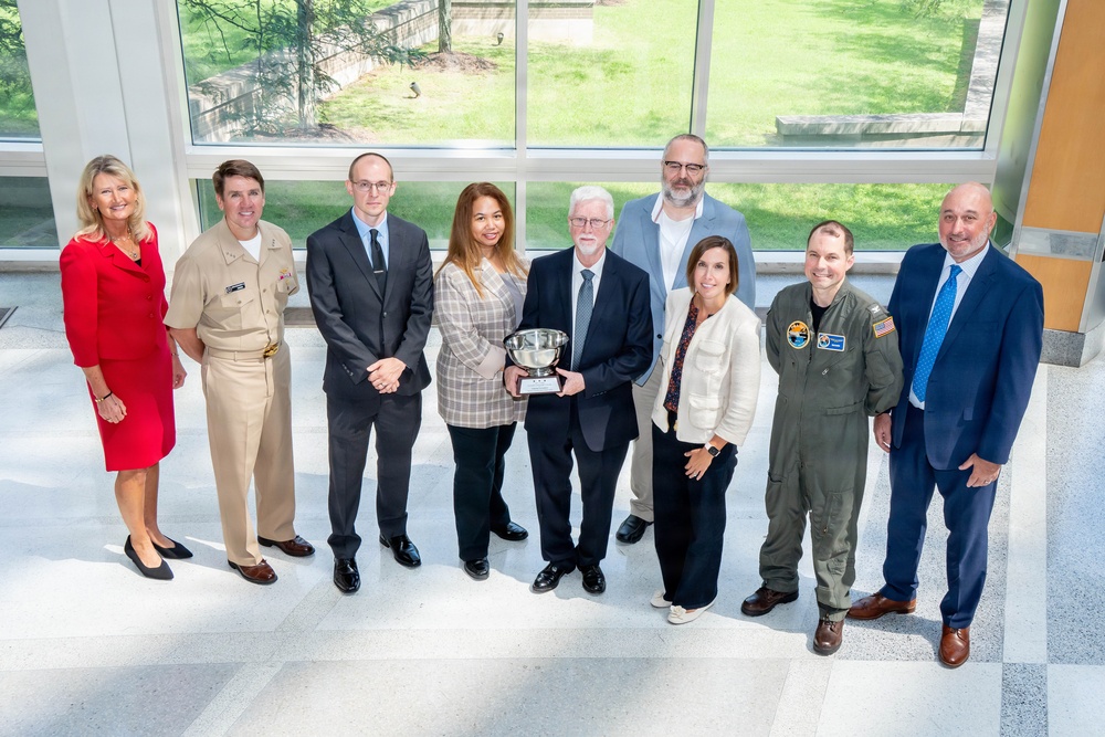 A group of nine individuals pose for a formal group photo in what appears to be a modern building with large windows. A central figure holds a silver trophy bowl, suggesting an award ceremony or recognition event. The group includes individuals in military uniforms, business attire, and flight suits.