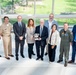 A group of nine individuals pose for a formal group photo in what appears to be a modern building with large windows. A central figure holds a silver trophy bowl, suggesting an award ceremony or recognition event. The group includes individuals in military uniforms, business attire, and flight suits.