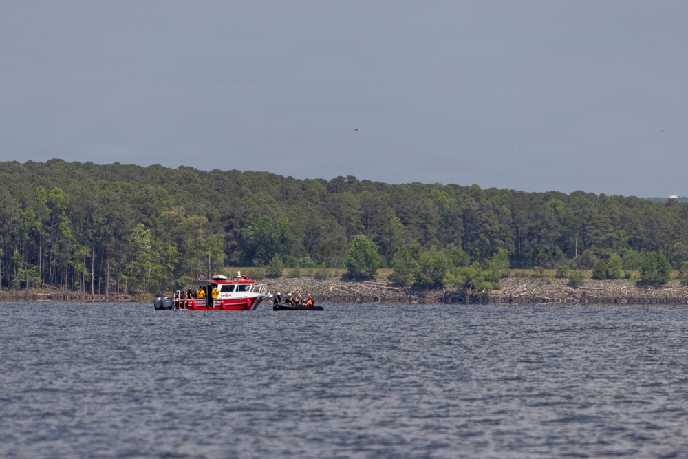 A red rescue or emergency response vessel is stationed on a large body of water, accompanied by what appears to be a smaller inflatable boat with personnel aboard. The scene shows multiple individuals engaged in what appears to be a water-based operation near a debris field along the shoreline. A dense treeline forms the background under an overcast sky.