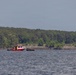 A red rescue or emergency response vessel is stationed on a large body of water, accompanied by what appears to be a smaller inflatable boat with personnel aboard. The scene shows multiple individuals engaged in what appears to be a water-based operation near a debris field along the shoreline. A dense treeline forms the background under an overcast sky.