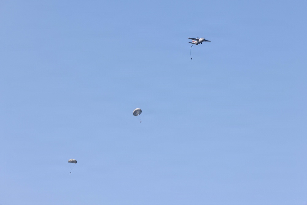 A military or training parachute jump operation is underway, with a twin-engine aircraft visible in the upper right portion of the image. Two parachutes are fully deployed and descending, while at least one jumper appears to have just exited the aircraft. The scene takes place against a clear, uniform blue sky with no clouds.
