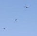 A military or training parachute jump operation is underway, with a twin-engine aircraft visible in the upper right portion of the image. Two parachutes are fully deployed and descending, while at least one jumper appears to have just exited the aircraft. The scene takes place against a clear, uniform blue sky with no clouds.