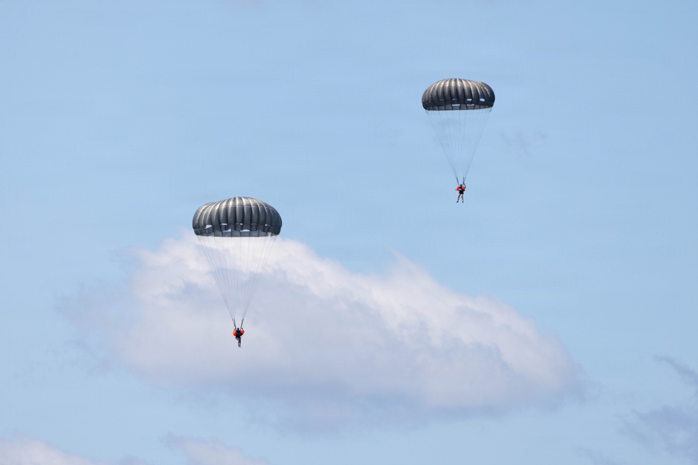 Two parachutists are descending through a clear blue sky with white clouds in the background. Both parachutes are dark gray/olive colored round canopies, consistent with military-style parachutes. Each jumper appears to be wearing bright orange gear or harness components, visible against the sky.