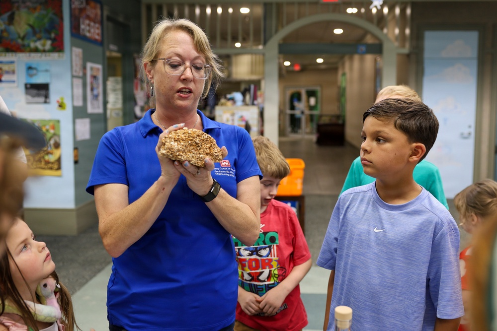 A woman wearing a blue polo shirt with a U.S. Army Corps of Engineers logo is holding and presenting a rock or mineral specimen to a group of children in what appears to be a museum or educational facility. The children, ranging in age from approximately 5 to 10 years old, are gathered around her attentively. The setting suggests an organized educational tour or outreach program.