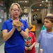 A woman wearing a blue polo shirt with a U.S. Army Corps of Engineers logo is holding and presenting a rock or mineral specimen to a group of children in what appears to be a museum or educational facility. The children, ranging in age from approximately 5 to 10 years old, are gathered around her attentively. The setting suggests an organized educational tour or outreach program.