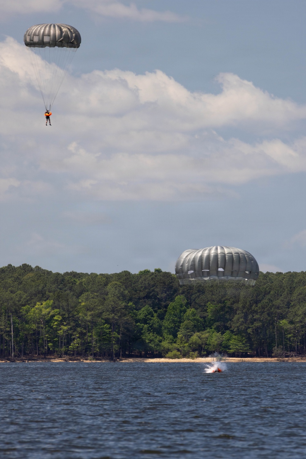 The image shows a military water jump training exercise over a lake surrounded by dense pine forest. Two parachutists with round, olive/gray military parachutes are visible — one still descending in the upper left, and another just entering the water with a visible splash in the lower center. The scene is consistent with airborne water landing training.
