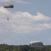 The image shows a military water jump training exercise over a lake surrounded by dense pine forest. Two parachutists with round, olive/gray military parachutes are visible — one still descending in the upper left, and another just entering the water with a visible splash in the lower center. The scene is consistent with airborne water landing training.