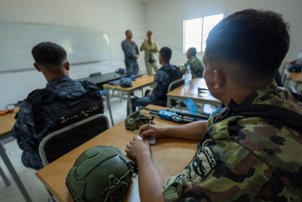 A military classroom training session showing personnel in various camouflage uniforms seated at desks with tactical equipment. Two instructors or senior personnel are standing at the front of the room near a whiteboard, appearing to conduct a briefing or instruction. Military gear including helmets, rifles, and tactical bags are visible on the desks.