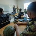 A military classroom training session showing personnel in various camouflage uniforms seated at desks with tactical equipment. Two instructors or senior personnel are standing at the front of the room near a whiteboard, appearing to conduct a briefing or instruction. Military gear including helmets, rifles, and tactical bags are visible on the desks.