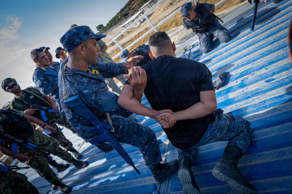 Military or law enforcement personnel are conducting a hands-on training exercise on blue-painted bleacher steps. A uniformed instructor in digital camouflage is demonstrating a control or restraint technique on a person in a black shirt. Multiple other uniformed personnel observe the demonstration, some carrying blue training rifles.