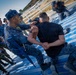 Military or law enforcement personnel are conducting a hands-on training exercise on blue-painted bleacher steps. A uniformed instructor in digital camouflage is demonstrating a control or restraint technique on a person in a black shirt. Multiple other uniformed personnel observe the demonstration, some carrying blue training rifles.