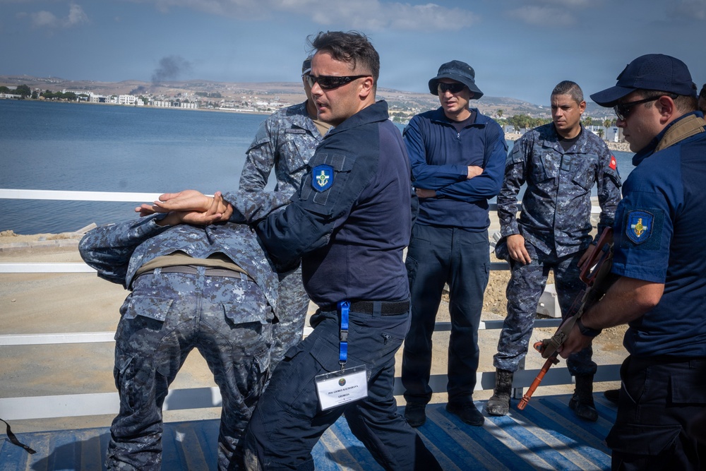 The image depicts what appears to be a military or law enforcement training exercise taking place on an outdoor platform near a waterfront. A central figure wearing a blue uniform with a lanyard/ID badge is demonstrating or practicing a control/restraint technique on another uniformed individual. Several other uniformed personnel are observing the demonstration.