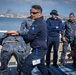 The image depicts what appears to be a military or law enforcement training exercise taking place on an outdoor platform near a waterfront. A central figure wearing a blue uniform with a lanyard/ID badge is demonstrating or practicing a control/restraint technique on another uniformed individual. Several other uniformed personnel are observing the demonstration.